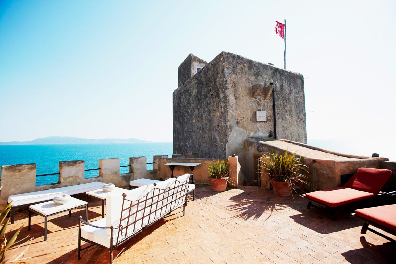 Historic stone tower with a flag overlooking a serene blue sea, surrounded by outdoor seating and potted plants, ideal for relaxation and scenic views. | Only on SopranoVillas