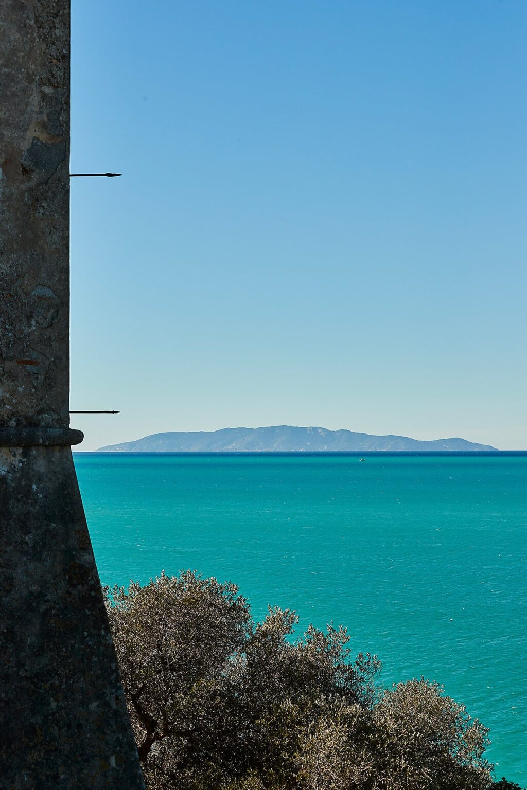 Scenic view of a distant island across a turquoise sea, framed by a stone structure on the left, under a clear blue sky. | Only on SopranoVillas