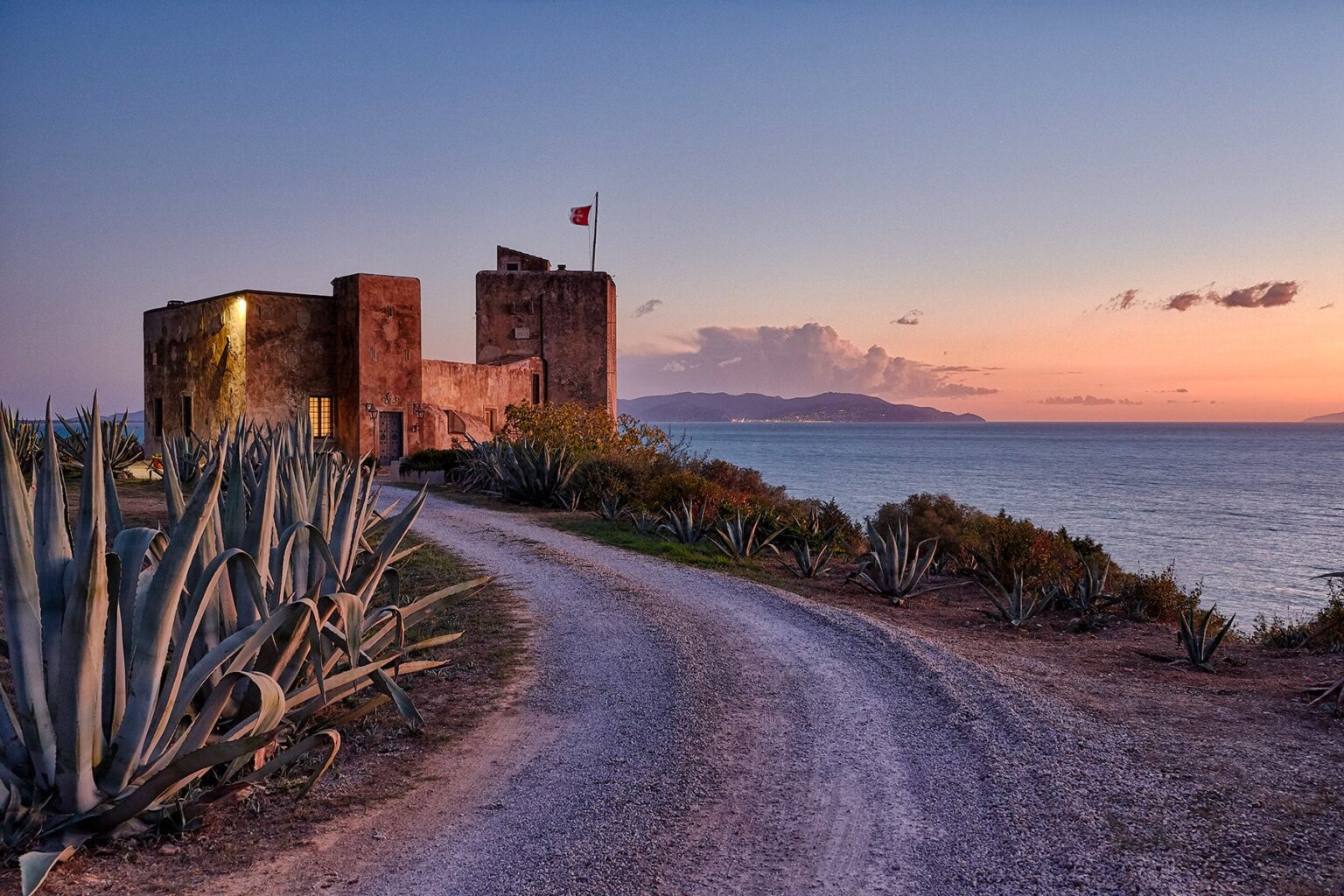 Historic coastal castle at sunset with a winding path and agave plants in the foreground. The scene captures the tranquil beauty of the seaside landscape and the architectural charm of the castle. | Only on SopranoVillas