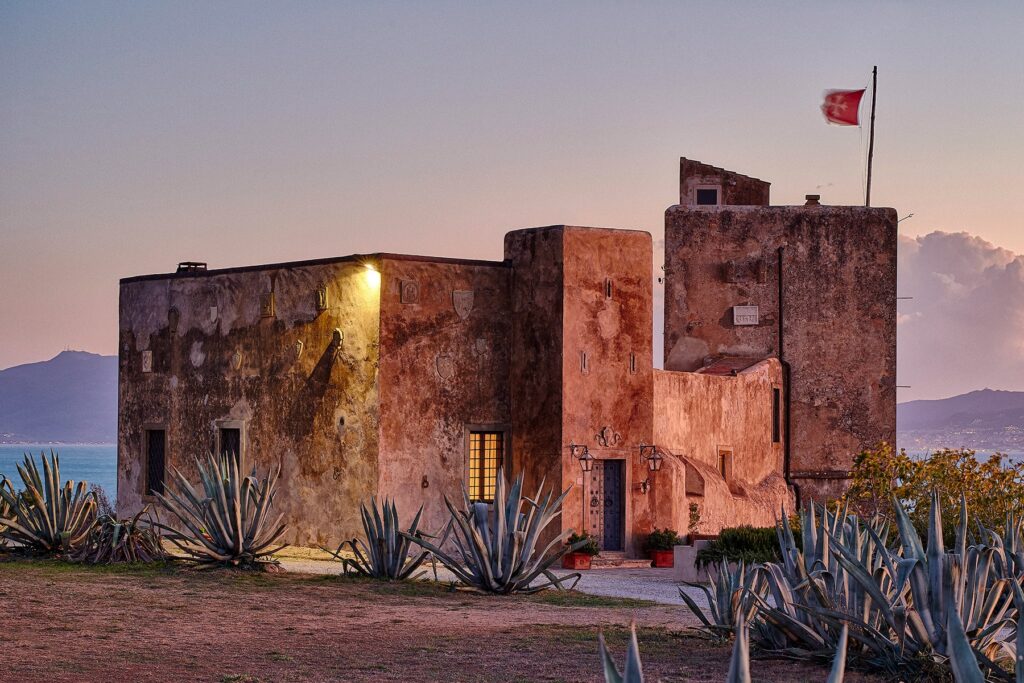 Historic stone building with a flag, surrounded by agave plants at sunset, showcasing coastal architecture and natural beauty. | Only on SopranoVillas