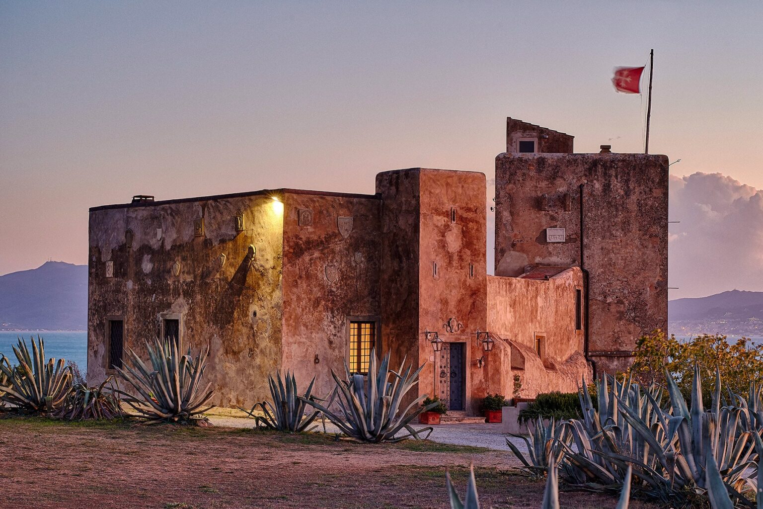 Historic stone building with a flag, surrounded by agave plants at sunset, showcasing coastal architecture and natural beauty. | Only on SopranoVillas