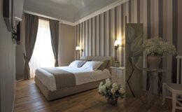 Modern bedroom interior featuring a stylish bed with neutral bedding, elegant striped walls, and soft lighting from bedside lamps. The room includes a decorative partition, a vase of flowers, and wooden flooring, creating a serene and inviting atmosphere.