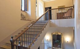 Interior view of a staircase in a well-lit hallway, featuring wooden railings, a decorative mirror, and a vintage chair, showcasing a blend of classic and contemporary design elements.