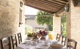 Outdoor dining setup featuring a rustic wooden table adorned with breakfast items, including pastries, fruits, and beverages, under a charming stone porch with natural light and greenery in the background.