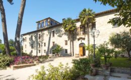 Historic stone building surrounded by lush greenery and colorful flowers, featuring large windows and an inviting entrance, ideal for a serene getaway or event venue.