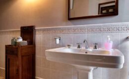 Modern bathroom featuring a white sink with chrome faucets, a wooden cabinet for storage, and a decorative mirror. The walls are tiled with a subtle pattern, and there are toiletries displayed for convenience.