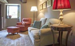 Cozy living room featuring a beige sofa, an orange armchair, a matching ottoman, and a stylish side table with a red lampshade, complemented by natural light from a window.
