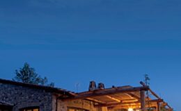 Cozy stone cottage illuminated at dusk, surrounded by trees under a clear blue sky, showcasing rustic architecture and warm ambiance.