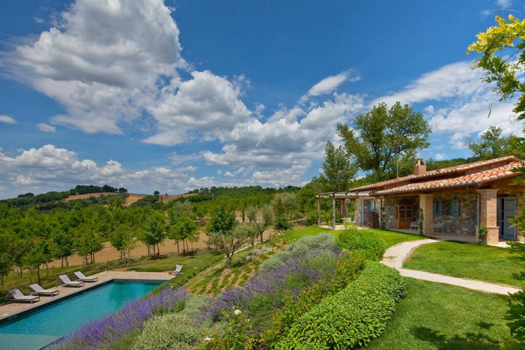 Scenic view of a luxurious villa with a swimming pool, surrounded by lush greenery and blooming flowers under a bright blue sky with fluffy clouds. Perfect for vacation rentals or outdoor events in a tranquil setting. | Only on SopranoVillas