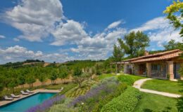 Scenic view of a luxurious villa with a swimming pool, surrounded by lush greenery and blooming flowers under a bright blue sky with fluffy clouds. Perfect for vacation rentals or outdoor events in a tranquil setting.
