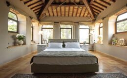 Spacious and elegant bedroom featuring a large bed with a stylish headboard, wooden ceiling beams, and ample natural light from large windows. Decorated with plants and tasteful furnishings, this serene space offers a calm and inviting atmosphere.