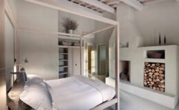 Modern bedroom interior featuring a four-poster bed, minimalist decor, and a cozy fireplace with stacked wood. The room has natural light, wooden beams, and neutral tones, creating a serene atmosphere.