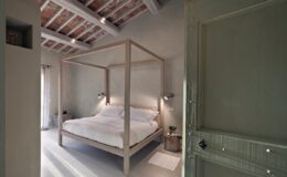 Modern bedroom featuring a four-poster bed with white linens, minimalistic decor, and natural light streaming through a window, highlighting the rustic ceiling beams and soft color palette.