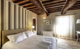 Cozy bedroom with wooden beams, striped walls, a large bed with white linens, and natural light streaming through sheer curtains.