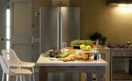 Modern kitchen interior featuring a stainless steel refrigerator, a wooden dining table with fresh vegetables, fruits, and herbs, and minimalist white chairs. The warm lighting enhances the inviting atmosphere of the space.