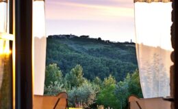 View from a window with sheer curtains, showcasing a picturesque landscape of rolling hills and lush greenery under a colorful sunset sky.
