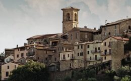Historic stone buildings of a medieval village with a bell tower, surrounded by greenery and a cloudy sky.