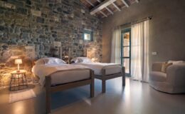 Cozy bedroom with two twin beds, stone walls, and large windows allowing natural light. Features a comfortable armchair and soft lighting for a relaxing atmosphere. Ideal for a tranquil getaway.