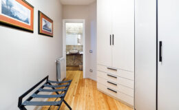 Modern hallway interior featuring sleek white cabinets, wooden flooring, and framed artwork on the walls, with a view into a bathroom at the end.