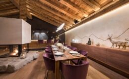 Modern dining room in a rustic chalet with wooden beams, featuring a long table set for a meal, purple chairs, and a cozy fireplace. The wall displays artistic mountain and animal motifs, creating a warm and inviting atmosphere.