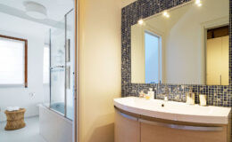 Modern bathroom featuring a sleek curved vanity with a blue mosaic backsplash, a large mirror, and a glass shower enclosure, complemented by natural light from a nearby window.
