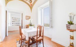 Bright and airy dining area featuring a wooden table with elegant chairs, a vase of orchids, and large windows that offer scenic views. The room is adorned with decorative elements, including a patterned rug and artistic ceiling details.