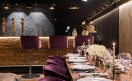 Elegant restaurant interior featuring a long wooden dining table set with fine china, glasses, and floral arrangements. Plush purple bar stools line the counter, illuminated by warm lighting, creating a cozy and sophisticated ambiance.