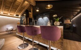 Modern kitchen interior featuring stylish purple bar stools, wooden accents, and ambient lighting. The design blends rustic and contemporary elements, creating an inviting atmosphere.