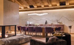 Modern dining room featuring a large wooden table surrounded by purple chairs, a cozy fireplace, and a minimalist mountain-themed wall design, illuminated by soft lighting for an inviting atmosphere.