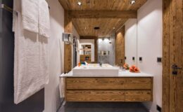 Modern bathroom featuring wooden accents, a sleek white sink, and contemporary lighting. The design includes a spacious vanity with storage, white towels hanging on the wall, and a minimalist aesthetic that enhances the overall ambiance.