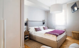 Cozy bedroom featuring a double bed with purple accents, natural light from a window, and modern decor elements, including a bedside table and a wall-mounted TV.