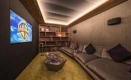 Cozy home theater featuring a large Warner Bros. screen, plush gray sofa with purple pillows, and modern shelving filled with books and decorative items, illuminated by soft ambient lighting.
