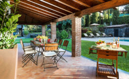Outdoor dining area under a wooden pergola, featuring a long table set for a meal, surrounded by lush greenery and lounge chairs in the background. Ideal for al fresco dining and gatherings.