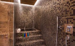 Modern spa shower featuring textured mosaic tiles, a built-in step, and colorful shampoo bottles arranged neatly. The ambient lighting enhances the serene atmosphere.