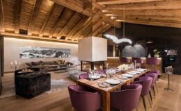 Modern dining room with a wooden ceiling, featuring a large table set for dinner with purple chairs, a contemporary fireplace, and stylish decor, creating a cozy and inviting atmosphere.
