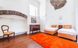 Cozy guest room featuring two twin beds, a wooden chair, and an orange rug, with bright white walls and a stone accent. Ideal for travelers seeking a comfortable and stylish accommodation.