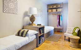 Cozy bedroom interior featuring two twin beds with white linens, a stylish lamp on a nightstand, and a small seating area with an accent chair. The room includes a closet with hanging clothes and decorative baskets, complemented by light wood flooring and wall art for a modern, inviting atmosphere.