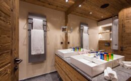 Modern bathroom interior featuring wooden accents, double vanity sinks, colorful glass accessories, and a spacious layout with natural lighting.