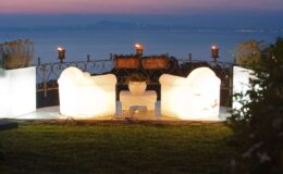 Illuminated outdoor seating area with two glowing chairs and a small table, overlooking a serene body of water at sunset, surrounded by lush greenery and decorative torches.