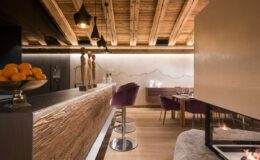 Modern restaurant interior featuring a wooden bar, stylish purple stools, and a cozy fireplace, with warm lighting and decorative mountain mural.