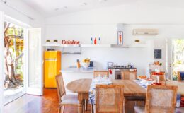 Bright and airy kitchen with a yellow retro refrigerator, wooden dining table, and modern appliances, featuring open doors leading to an outdoor area, designed for a cozy dining experience.