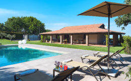 Modern villa with a swimming pool and lounge chairs under umbrellas, surrounded by lush greenery and a clear blue sky. Ideal for relaxation and outdoor gatherings.