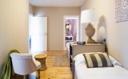 Cozy bedroom featuring a single bed with decorative pillows, a stylish chair, a small wooden stool, and a lamp on a bedside table. The room has warm wooden flooring and soft beige walls, creating a serene atmosphere. A door leads to another room, enhancing the spacious feel.