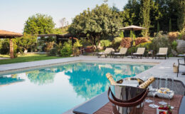Luxurious outdoor swimming pool surrounded by lush greenery, sun loungers, and shaded areas, with a stylish bucket of champagne and snacks on a table nearby, ideal for relaxation and leisure.