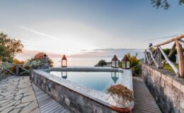 Scenic view of a tranquil swimming pool surrounded by natural landscape, with lanterns and a stone pathway, capturing a serene sunset over the water.