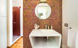 Modern bathroom featuring a sleek white sink with a unique geometric design, complemented by a vibrant, colorful mosaic wall and a round mirror. The space includes a shower area and wooden flooring, creating a contemporary and inviting atmosphere.
