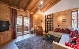 Cozy living room in a mountain chalet featuring wooden beams, a comfortable sofa with colorful cushions, a plush armchair, a coffee table, and a bookshelf, with large windows offering scenic views and natural light.