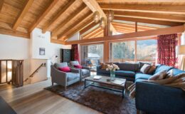 Cozy living room with high wooden ceilings, large windows showcasing mountain views, and stylish furniture including a blue sofa and gray armchairs, accented by colorful cushions and a decorative rug.