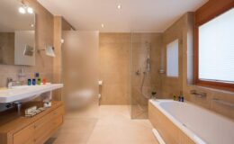 Modern bathroom interior featuring a glass shower, bathtub, wooden vanity with sink, and stylish decor. Natural light enters through a window, enhancing the spacious and elegant design.