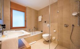Modern bathroom featuring a glass shower, bathtub, toilet, and natural light from a window, designed with neutral tile and wooden accents.